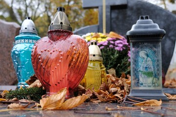 Colorful glass candles among dry autumn leaves on grave in cemetery on November day
