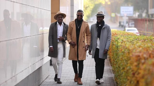 Three stylish African friends in hats walks together and talks in a city street
