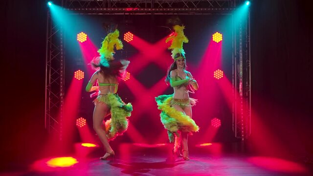 Two passionate young women dancing samba music at a carnival party. Dancers in traditional costumes and feather headdresses perform in the studio with red lights in the background. Slow motion.
