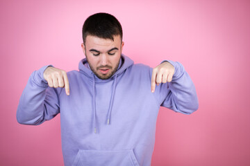 Young handsome man wearing casual sweatshirt over isolated pink background surprised, looking down and pointing down with fingers and raised arms