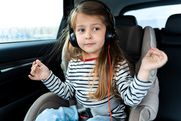 Little cute caucasian girl is driving in car.Kid listening to music with big headphones.Traveling,riding on road in safe baby seat with child belt.Fun family trip,entertainment, activity with parents