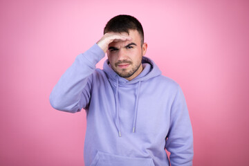 Young handsome man wearing casual sweatshirt over isolated pink background very happy and smiling looking far away with hand over head. searching concept.