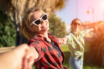 Pretty young caucasian woman in sunglasses pulling along by the hand invitingly with a happy smile as her friend on the background. Sunny summer day