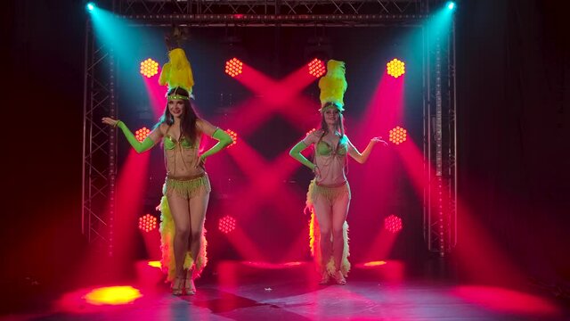 Two passionate young women dancing samba music at a carnival party. Dancers in traditional costumes and feather headdresses perform in the studio with red lights in the background. Slow motion.