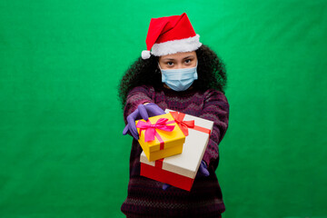Young woman in protective coronavirus mask and Santa hat holds Christmas gift boxes on green background.