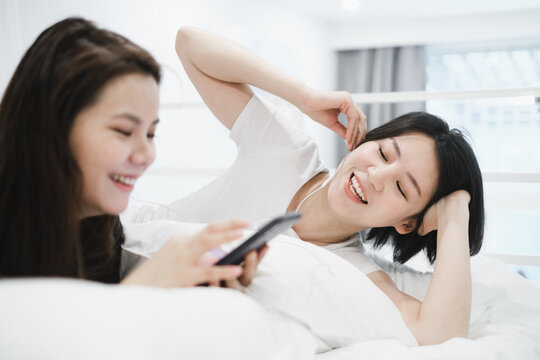 Two Beautiful Young Girlfriends Listening To Music With Cell Phone On Bed At Home