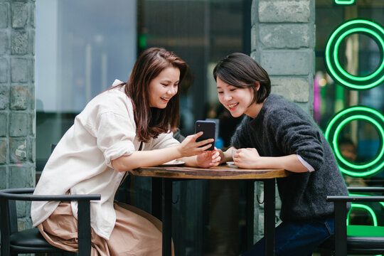 Young Women Friends Enjoying Coffee And Looking At Cell Phone In The Cafeteria