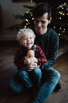 toddler (little boy) holding a doll and sitting on his fathers lap in front of the christmas tree with christmas lights