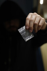 The hand of a hooded drug dealer, as he holds out a transparent sachet in front of him, with some contraband pills inside.