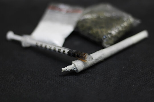 A syringe full of cocaine, placed on a cane of marjiuana or ganja, with a sachet full of cocaine, and a sachet full of ganja or marijuana placed in the background