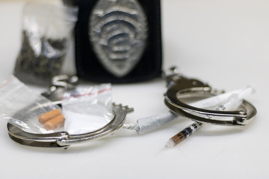 A Police-seized Drug Shipment Consisting Of Illegal Drugs, Cocaine Powder And Ganja Sachets, And A Syringe Full Of Cocaine Next To A Joint, Both In Handcuffs