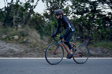 Motion blur of bearded cyclist wearing black windbreaker jacket with blue side, helmet, goggles training with bike on asphalt road. Strong man improving skills and getting ready for competition