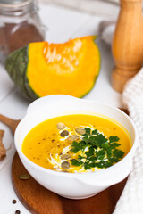 A plate of yellow pumpkin cream soup with cream, herbs and seeds on a white table near raw pumpkin and a wooden spoon.