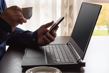 man in blue shirt drinking a cup of coffee while checking his cell phone and working from home with his laptop