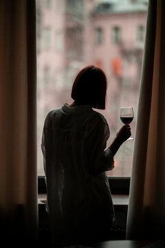 silhouette of a young girl in a white shirt with short hair, stands near the window with a glass of wine