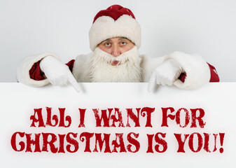 Santa Claus points his fingers at the board with the text - ALL I WANT FOR CHRISTMAS IS YOU