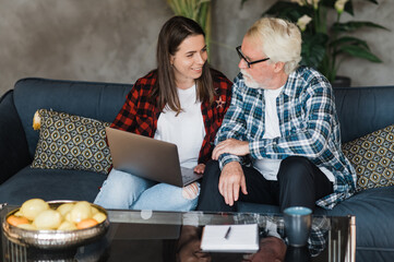 Young adult daughter and grey-haired father are sitting on the couch at home. The girl teaches her old dad to use laptop and social media