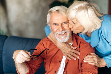 Happy elderly gray-haired couple hugging and sits on the sofa at home, wife kisses husband and...