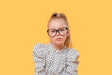 Portrait of a blonde Girl child with glasses for vision looking at the camera. Black-rimmed glasses. Yellow isolated background.