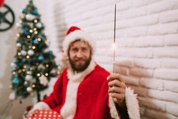 A young caucasian man in a Santa Claus costume holds a sprinkler in his hand while a Christmas tree...