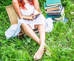 No face girl in spring blooming park Hand holding old retro open book Woman sittting in vintage aged suitcase on green grass against fresh green grass background near stack