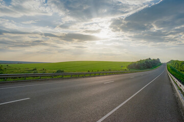 Empty asphalt road at sunset. Cloudy sky and green meadows.