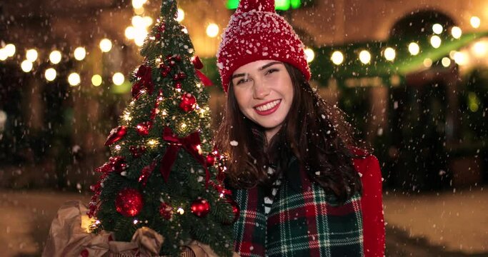 Close up portrait of happy Caucasian young woman in good mood standing on snowy street with small christmas tree, looking at camera and smiling Grateful female in decorated town at night New year mood