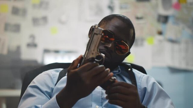 Confident african policeman in glasses sitting at desk with gun at police station