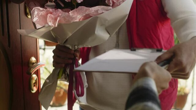 Tilt up shot of handsome middle eastern delivery man in uniform and cap smiling, taking signed document and giving bouquet of flowers to woman