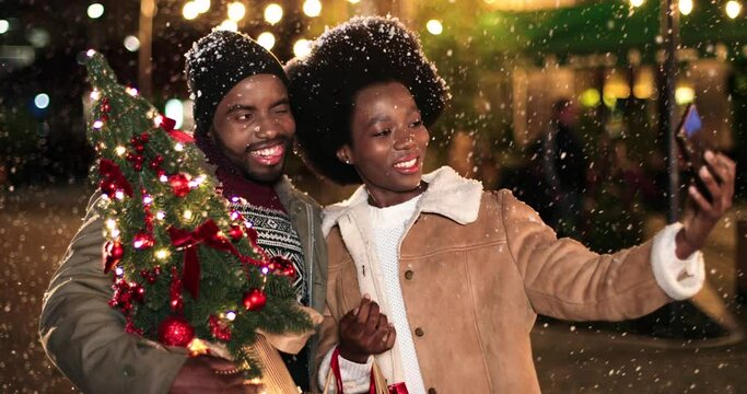 Portrait of happy smiling African American couple taking funny selfie photos on mobile phone on street with little christmas tree. Man and woman taking pictures at night on new years eve. Magic time