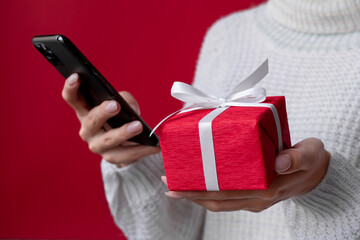 Christmas online shopping. Female hands holding smartphone and red gift box on the red background. White sweater. December sales and discounts promotions during winter holidays. Surprise delivery