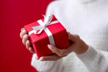 Female hands holding red gift box with white ribbon and bow on the red background. Christmas and New Year holidays. Valentine's Day and Birthday. Surprise, greeting, celebration, present. Isolated