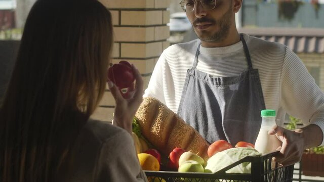 Cheerful male farmer in apron and cap smiling and giving crate with fresh produce, bread and milk to female customer while doing farm-to-door delivery service