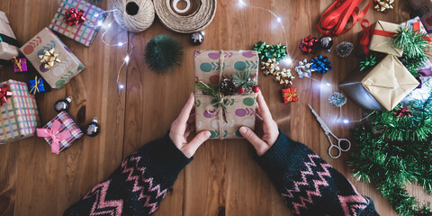 Young lady dressed in sweater holding christmas concept gift package in hand on table. Young lady's hand preparing stylish gift boxes.