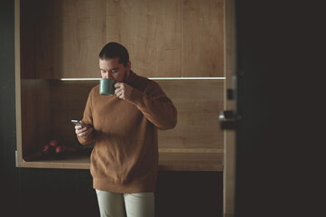 Young man drinks coffee and looks into a smartphone in the kitchen