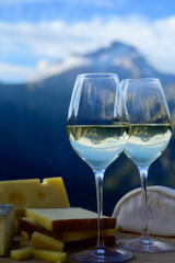 Tasty cheese and wine from Savoy region in France, beaufort, abondance, emmental, tomme and reblochon de savoie cheeses and glass of white wine served outdoor with Alpine mountains peaks on background