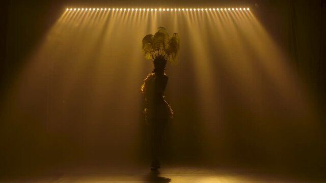 Silhouettes of a beautiful carnival dancer in a yellow suit and a headdress made of feathers. Gorgeous woman passionately dancing samba in the studio against the background of lights in slow motion.