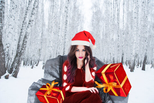 Portrait Young Latin Hispanic Brunette Sexy Santa Woman Wearing Red Christmas New Year Hat  Against Gift Box Winter Forest And Snow Drift Background Cute Girl Looking At Camera Black Princess Throne