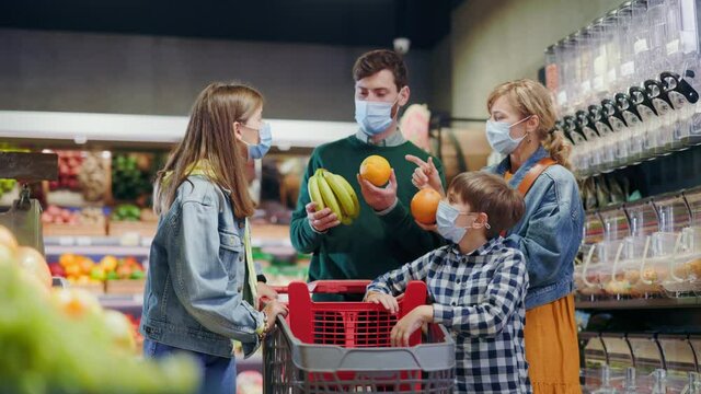 Quarantine. Caucasian young affectionate family of parents and kids selecting best quality foods natural fruits at grocery store. Supermarket. Weekend. Medical masks.