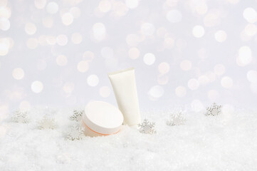 Set of winter skin care cosmetic products in snow and snowflakes on white background with bokeh lights. Face cream jar and hand creme or body lotion tube. Light festive composition with copy space