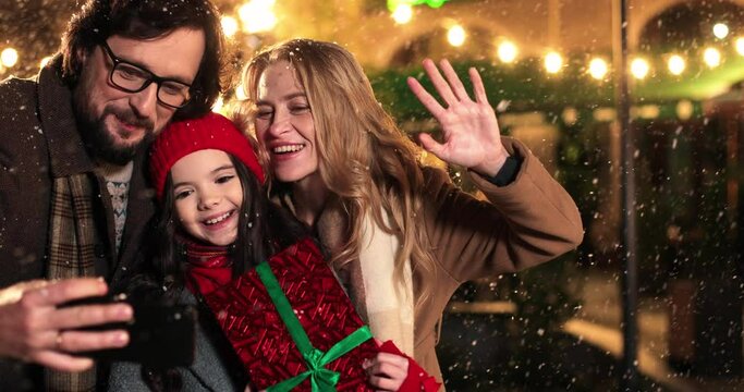 Portrait of happy Caucasian family taking pictures together on street while snowing on new years eve. Father, mother and cute daughter taking selfie photos on cellphone in decorated xmas city