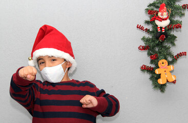 
7-year-old latin boy with protection mask, sweater and Christmas hat on gray background with...