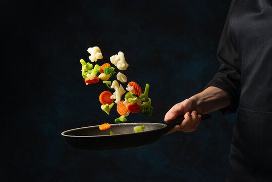 Professional Chef Hand In Black Uniform Throws Up Frying Mix Of Cabbage And Carrot Above The Pan On Dark Blue Background. Backstage Of Cooking Meal. Frozen Motion. Food Banner Concept.