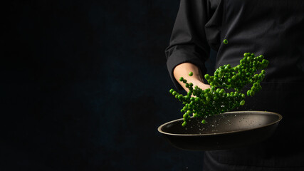 Professional chef in black uniform throws up frying green peas above the pan on dark blue background. Backstage of cooking meal. Frozen motion. Food banner concept.