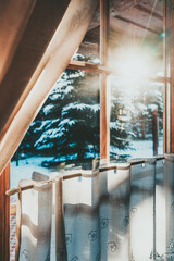 Bright Sunlight Shines Through the Wooden Cottage House Window. Beautiful snowy winter morning behind the chalet window. Sunshine and sparkling snow on fir trees in the cabin backyard