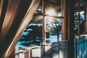 Bright Sunlight Shines Through the Wooden Cottage House Window. Beautiful snowy winter morning behind the chalet window. Sunshine and sparkling snow on fir trees in the cabin backyard