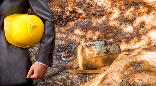 Hand or arm of engineer hold yellow plastic helmet for workers security on dry trash  hole in brown yellow toxic land with rusty old metal oil barrel and wooden board Ecology idea concept