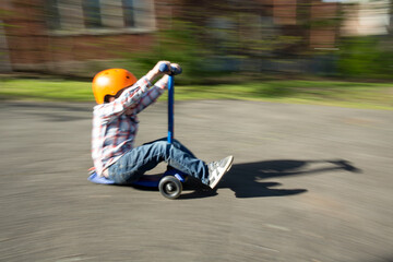Boy outside wearing orange helmet speeds down driveway on blue scooter