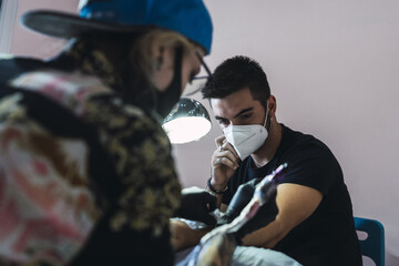 Man wearing a face mask and talking on phone when getting his hand tattooed in a tattoo salon