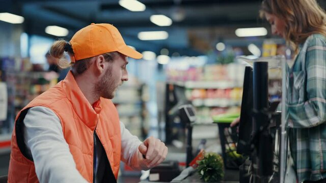 Young woman putting food items on cashier line shopping grocery store. Bearded attractive polite cashier worker serving the customer. Supermarket concept.